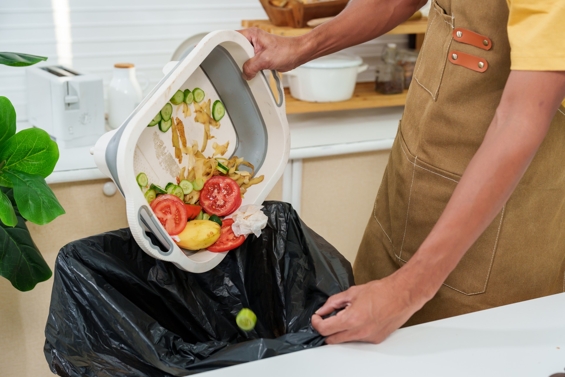 Close-up shot of a man's hand carrying food scraps and unused vegetable scraps Throw it in the trash with black bags. to separate different types of waste Wearing an apron in kitchen. stay home alone